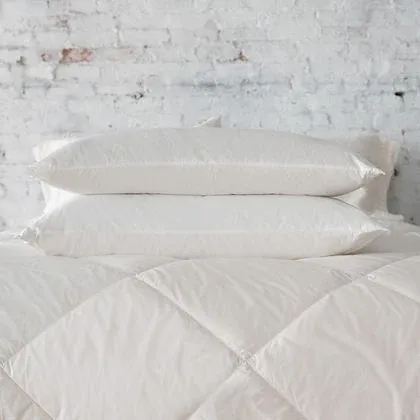 Three St. Genève Piatra Merino wool pillows stacked on a quilted bed against a white brick wall