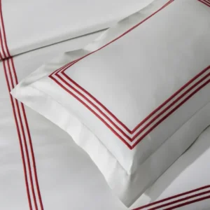 Close-up of elegantly styled white bedding with red striped accents, featuring a decorative pillow with a matching red border. Featuring Bel Tempo Embroidered bedding by Matouk. Available at Linen Society.