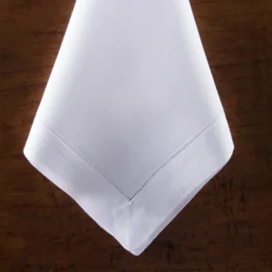 The Heirloom Estate Dinner Napkin by Henry Handwork, crafted from white Italian linen with a delicate hemstitch border, is folded and displayed on a dark wooden surface.