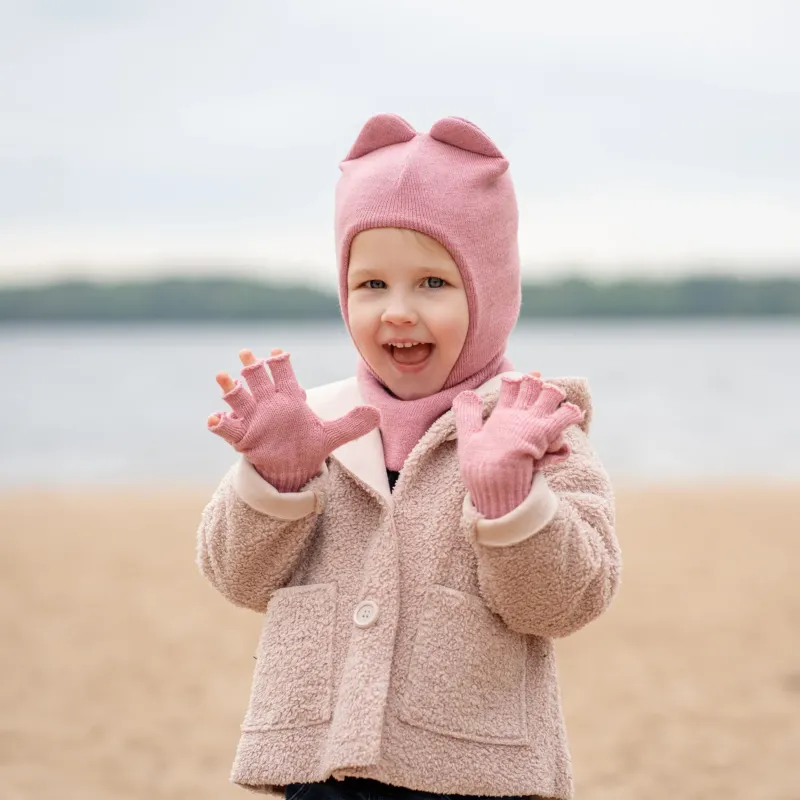 A joyful child wearing a pink balaclava with ears, a pink scarf, and a beige coat stands on a beach, hands open in excitement.