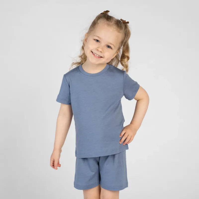A young child poses playfully with one hand on their hip and a bright smile, wearing a dusty blue short-sleeve t-shirt and matching shorts. They have blonde hair styled in half-up pigtails with small brown clips, set against a plain light background.
