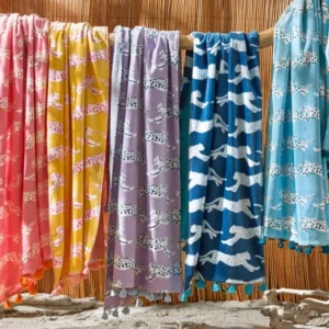 Five colorful Schumacher Leaping Leopard beach towels by Matouk hang on a wooden pole against bamboo, with sand and driftwood in front—ideal for poolside lounging or as your next standout beach towel.