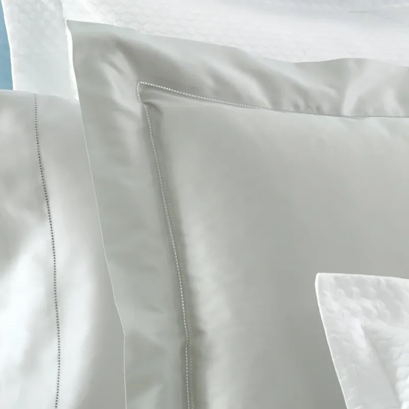 A close-up of elegant, smooth bedding featuring soft gray and white pillows with decorative stitching and intricate patterns. Featuring Nocturne Hemstitch bedding by Matouk, available at Linen Society.